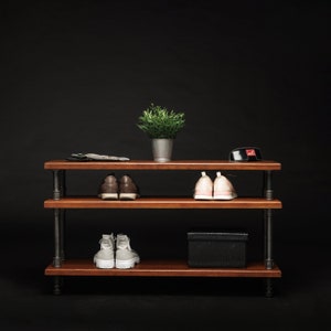 May include: A three-tiered wooden shelf unit with black metal pipes for legs. The shelves are filled with shoes, a small potted plant, and a black box.