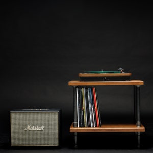 May include: A black Marshall Bluetooth speaker sits on a black surface next to a wooden record player stand with a turntable and a stack of vinyl records. The stand has black metal legs.