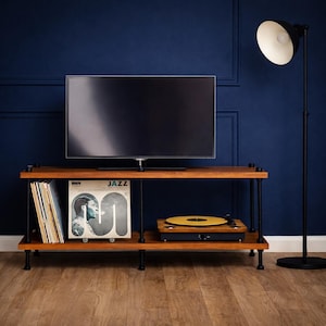 May include: A wooden media unit with a black frame, holding a flat-screen TV, vinyl records, and a record player. The unit is set against a dark blue wall with a black floor lamp. The record player has a yellow record on it.