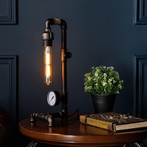 May include: Industrial-style table lamp made from black metal pipes, featuring a vintage Edison bulb. The lamp includes a pressure gauge and sits on a round wooden table. A small potted plant and a leather-bound book are also visible.