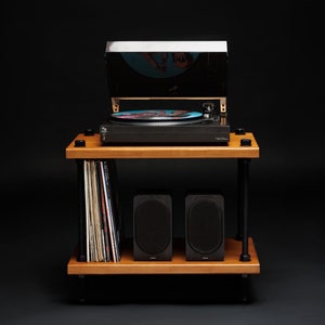 May include: A wooden record player stand with a black turntable, a stack of vinyl records, and two black speakers. The stand has two shelves and black metal supports. The turntable has a clear cover and a record is playing.
