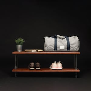 May include: A two-tiered wooden shelf with black metal pipe legs. A grey duffel bag sits on the top shelf, with two pairs of shoes on the bottom shelf. A small potted plant and two books are also on the top shelf.
