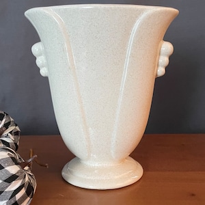 Puede incluir: Un jarrón de cerámica blanco con un acabado texturizado y lados acanalados. El jarrón tiene dos asas a los lados y una base de pedestal.