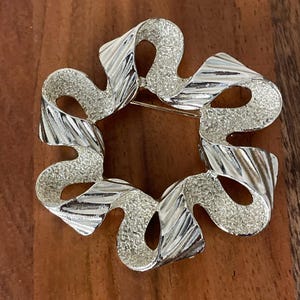 May include: A silver-toned brooch in the shape of a stylised ribbon bow. The bow is textured and has a slightly raised design.