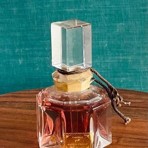 May include: A vintage perfume bottle with a clear, rectangular stopper and a faceted, octagonal body. The bottle contains a reddish-brown liquid and has a gold label that reads "FABERGE" and "MADE IN FRANCE". A brown cord is tied around the neck.
