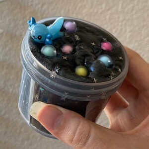 May include: A clear plastic container filled with black slime with glitter and colorful balls. A blue and white cartoon character is on top of the slime.