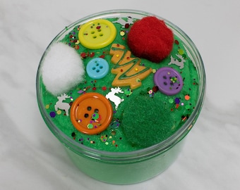 May include: A green slime with various Christmas-themed decorations, including a red pom-pom, a white cotton ball, a yellow button, a blue button, a purple button, an orange button, a gold Christmas tree, and silver reindeer.
