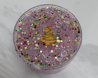 May include: A clear plastic container filled with pink slime, glitter, and clear beads. A small gingerbread cookie shaped like a Christmas tree is on top of the slime.