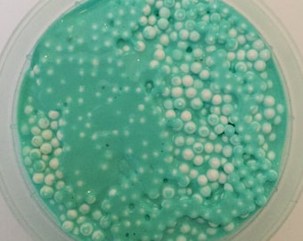 May include: A close-up of a teal blue slime with white foam balls in a clear plastic container.