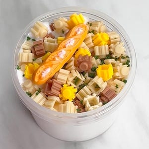 May include: A clear plastic container filled with white slime, topped with miniature food-themed decorations. The decorations include small, colorful blocks, green sprinkles, and a miniature baguette. The slime has a smooth, creamy texture.
