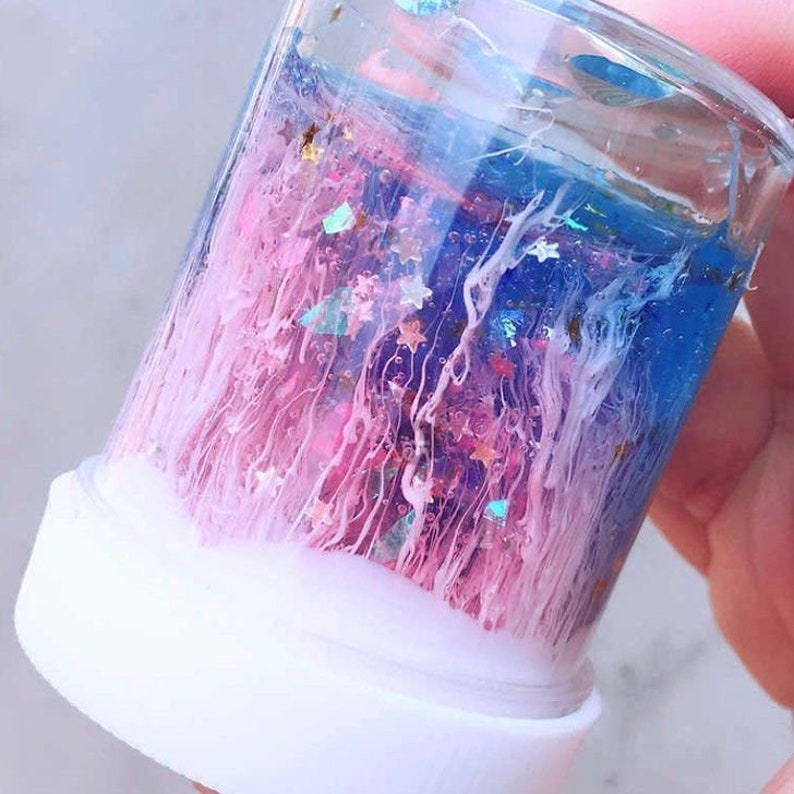 May include: A clear glass jar with a white base filled with a blue and pink liquid with glitter and white swirls.