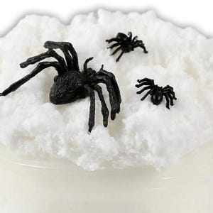 May include: A jar filled with white, fluffy material, possibly slime or a similar substance, topped with three black plastic spiders. The spiders vary in size, with one large spider and two smaller ones.