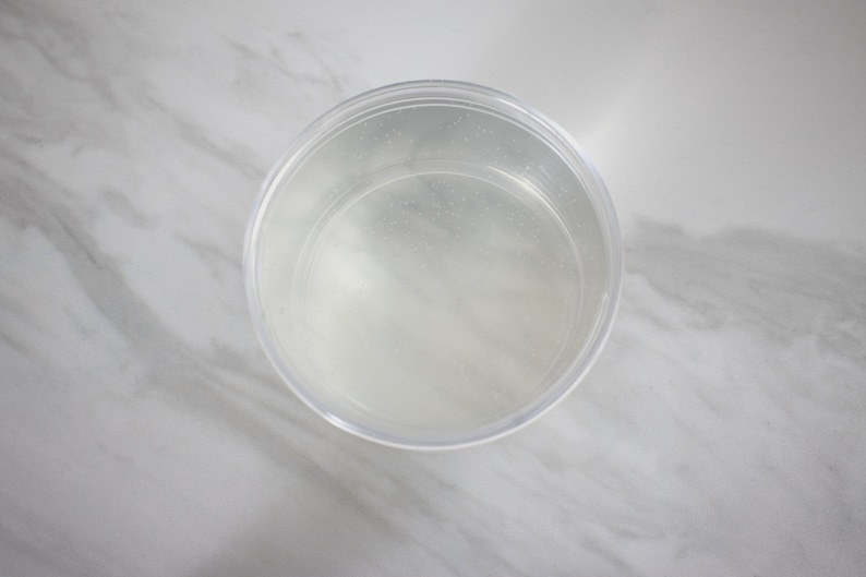Op de afbeelding: Een doorzichtige plastic container met een gladde, ronde bodem en een licht verhoogde rand. De container is gevuld met een heldere vloeistof.