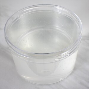 Op de afbeelding: Een doorzichtige plastic container met een deksel, gevuld met een heldere vloeistof.