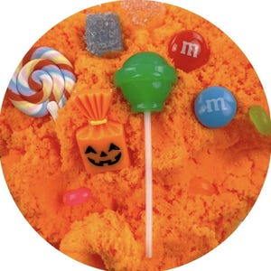 May include: A circular arrangement of orange-colored sand, adorned with various candies. A green lollipop, a swirl lollipop, and a small orange bag with a jack-o'-lantern face are visible. Red and blue candies with the letter "m" are also present.