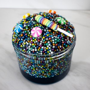 Può includere: Un barattolo di vetro trasparente pieno di slime blu con spruzzi colorati e decorazioni di caramelle in cima. Le caramelle includono un lecca-lecca arcobaleno, una caramella alla menta verde e bianca e una caramella alla menta blu e bianca.