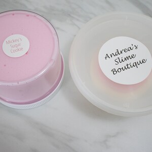 May include: Two clear plastic containers with pink slime inside. The container on the left has a white label that says "Mickey's Sugar Cookie". The container on the right has a white label that says "Andrea's Slime Boutique".
