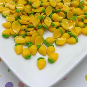 May include: A large pile of yellow and green mango-shaped resin charms. The charms are scattered on a white surface.