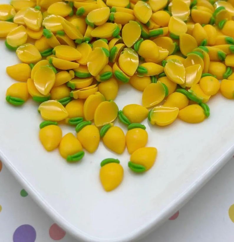 May include: A large pile of yellow and green mango-shaped resin charms. The charms are scattered on a white surface.