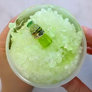 May include: A clear plastic cup filled with green slime and small white beads. A miniature green bottle with the word "Aloe" printed on it is partially submerged in the slime.