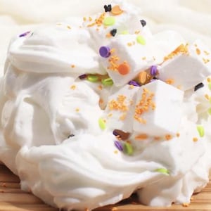 May include: A close-up of a white, fluffy substance, possibly slime or frosting, adorned with colorful sprinkles and square marshmallows. The sprinkles are orange, green, purple, and black, creating a festive appearance.