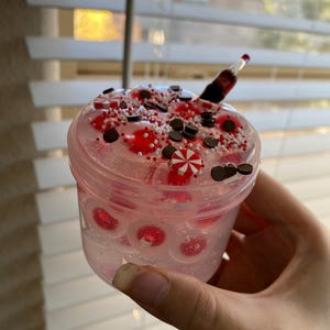 Puede incluir: Un recipiente de plástico transparente lleno de slime rosa, caramelos rojos y blancos, chispas de chocolate y espolvorear blanco. Una botella en miniatura de rayas rojas y blancas sobresale del slime.