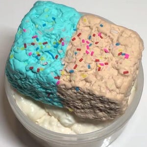 May include: A clear plastic container filled with slime. The top is split into two textured sections: one light blue, the other tan, both scattered with colourful sprinkles. The slime sits on a white, creamy base.