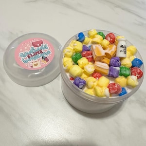 Puede incluir: Un recipiente de plástico transparente lleno de una sustancia viscosa blanca. La slime está cubierta con piezas de cereal de colores, malvaviscos y un pequeño amuleto de cartón de leche. El recipiente tiene una tapa rosa con el texto "Andrea's Slime Boutique" impreso en ella.