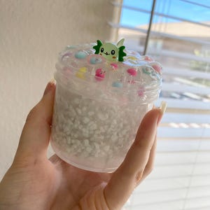 May include: A clear plastic container filled with white pebbles and clear slime. The slime is decorated with colorful beads and a green cartoon character. The container is held in a hand.