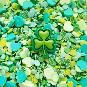 May include: A close-up of green, blue, and yellow sprinkles in the shape of hearts, shamrocks, and circles. A large green shamrock sprinkle is in the center of the image.