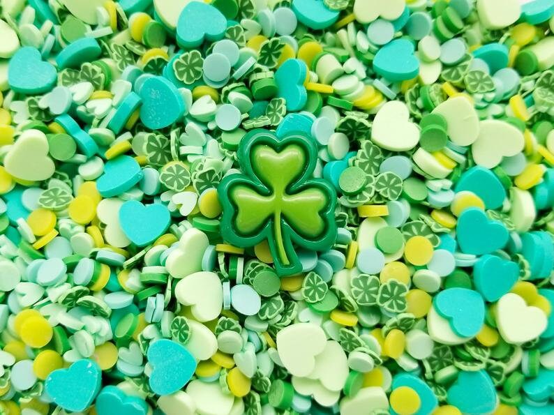 May include: A close-up of green, blue, and yellow sprinkles in the shape of hearts, shamrocks, and circles. A large green shamrock sprinkle is in the center of the image.