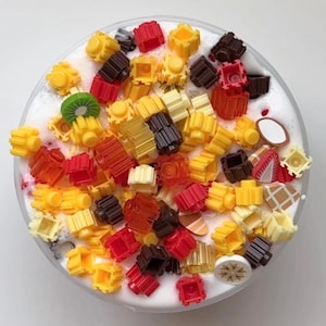 May include: A clear container filled with white slime and colorful, decorative pieces. The pieces are various shapes and colors, including yellow, red, brown, and clear. There are also slices of fruit and other decorative elements.