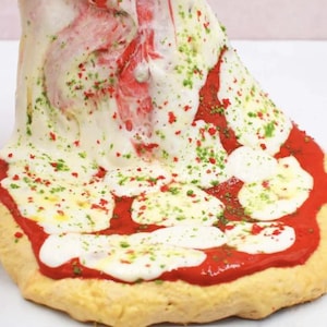 May include: A close-up of a pizza slice with melted white cheese, red sauce, and green and red sprinkles. The crust is golden brown. The cheese is stretching upwards, creating a visually appealing effect.