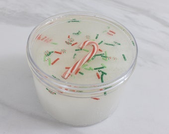 May include: A clear plastic container filled with white slime with red and green sprinkles and a candy cane on top.