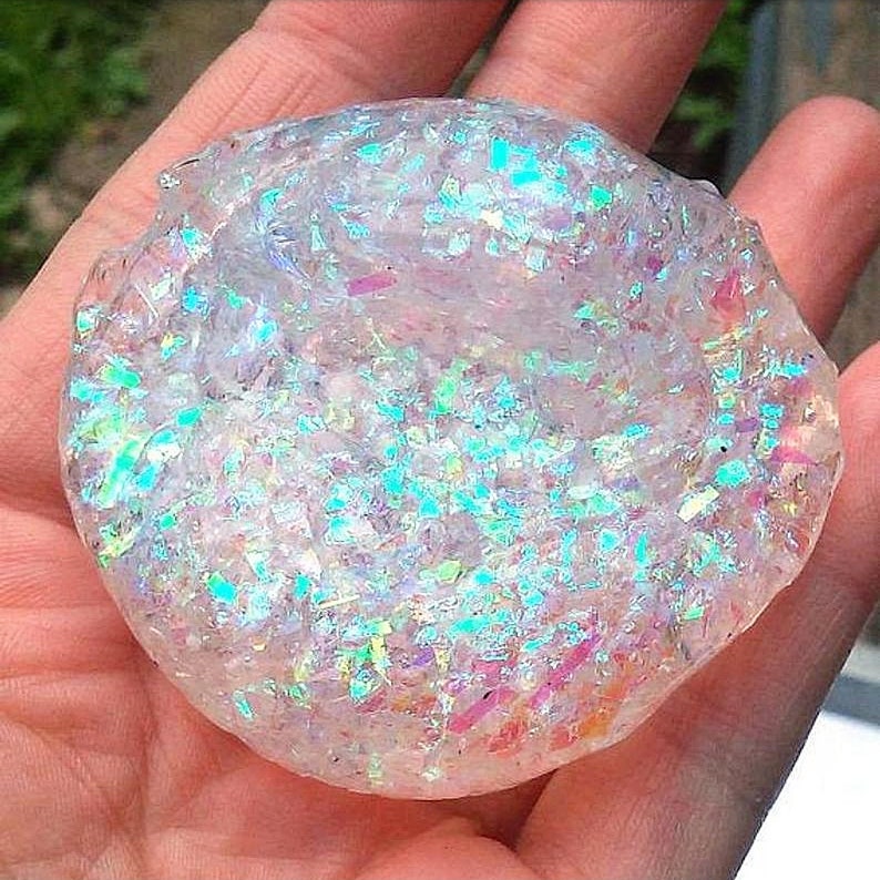 May include: A clear, round object filled with iridescent glitter, held in a hand. The glitter reflects shades of blue, green, pink, and purple, creating a shimmering effect. The object appears to be a decorative item or craft supply.