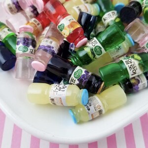 May include: A collection of miniature plastic bottles with various colors and designs. The bottles have Korean text on them and some have fruit designs. The bottles are on a white plate with pink and white stripes.