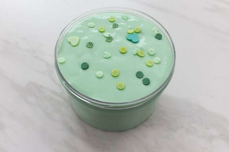 May include: A clear container filled with light green slime with various green, yellow, and blue sprinkles.