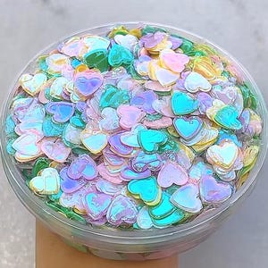 May include: A clear plastic container filled with iridescent heart-shaped confetti in shades of pink, blue, green, and yellow. The confetti has a shiny, reflective surface, creating a colorful and playful effect. The container is held by a person.