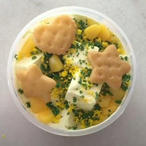 May include: A clear plastic container filled with a creamy white substance topped with yellow corn kernels, green sprinkles, and various shaped biscuits.