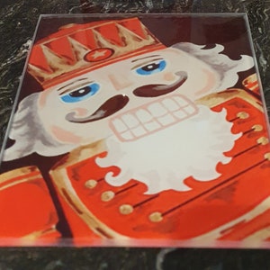 May include: A close-up of a nutcracker figurine with a red and gold uniform, a white beard, and a blue eye. The nutcracker has a star on its hat and a moustache.