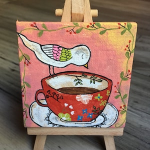 May include: A small painting on canvas of a white bird with pink and green accents perched on the rim of a red teacup with a floral pattern. The teacup is on a white saucer with a black lace trim. The painting is on a small wooden easel.
