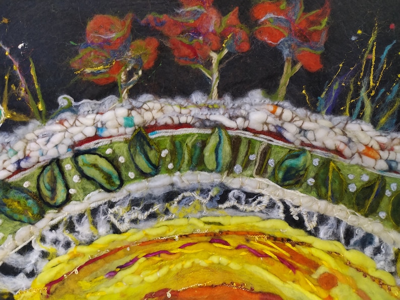 Op de afbeelding: Een close-up van een viltkunstwerk dat een zonsondergang over een landschap voorstelt. De lucht is diep zwart met rode, oranje en gele bomen. De voorgrond is een groen veld met witte en groene bladeren. De zon is helder geel en oranje.
