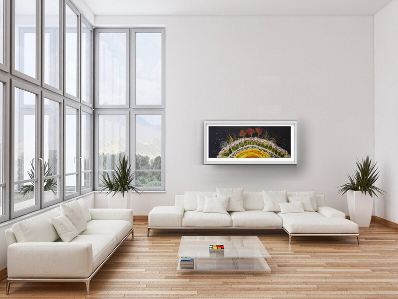 Op de afbeelding: Een ingelijst abstract schilderij in geel, oranje en groene tinten. Het schilderij toont een cirkelvormige vorm met een donkere achtergrond en een helder centrum. Het schilderij hangt aan een witte muur in een woonkamer.