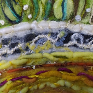Op de afbeelding: Close-up van een kleurrijke, textuurrijke, ronde wandhanger gemaakt van wolgaren en borduurwerk. De hanger heeft een verscheidenheid aan kleuren, waaronder groen, geel, oranje en paars. De bovenkant van de hanger is versierd met een witte rand en een laag groene en bruine wol.