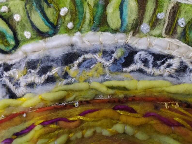 Op de afbeelding: Close-up van een kleurrijke, textuurrijke, ronde wandhanger gemaakt van wolgaren en borduurwerk. De hanger heeft een verscheidenheid aan kleuren, waaronder groen, geel, oranje en paars. De bovenkant van de hanger is versierd met een witte rand en een laag groene en bruine wol.