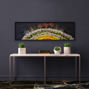 Op de afbeelding: Een lang, horizontaal schilderij met een zwarte achtergrond en een kleurrijk, abstract ontwerp. Het schilderij toont een gele, oranje en rode zonachtige vorm met groene bladeren en rode bloemen. Het schilderij is in zwart ingelijst en hangt aan een donkergrijze muur boven een witte marmeren consoletafel met twee potplanten.