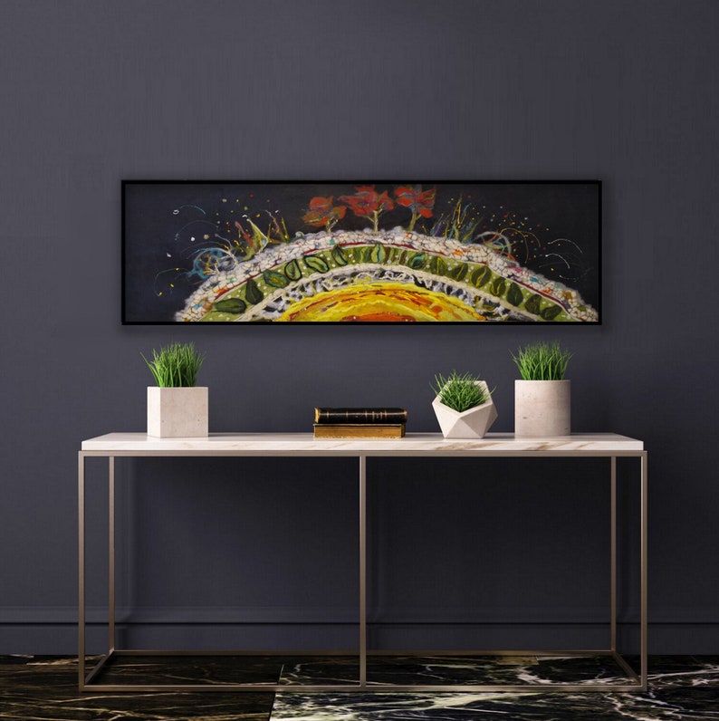 Op de afbeelding: Een lang, horizontaal schilderij met een zwarte achtergrond en een kleurrijk, abstract ontwerp. Het schilderij toont een gele, oranje en rode zonachtige vorm met groene bladeren en rode bloemen. Het schilderij is in zwart ingelijst en hangt aan een donkergrijze muur boven een witte marmeren consoletafel met twee potplanten.