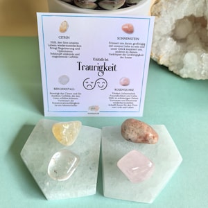 Könnte beinhalten: Vier kleine Kristalle auf weißen Sockeln, beschriftet mit ihren Namen und Eigenschaften. Die Kristalle sind Citrin, Sunstone, Bergkristall und Rosenquarz. Der Text auf der Karte lautet "Kristalle bei Traurigkeit".