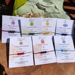Op de afbeelding: Zeven kleurrijke kaarten met chakra-symbolen, namen en beschrijvingen. Elke kaart heeft een armband van touw met kralen die de chakra vertegenwoordigen. De kaarten zijn in een rij op een groene ondergrond gerangschikt.