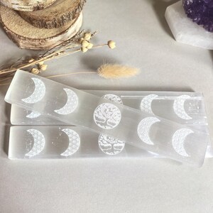 Può includere: Diverse bacchette rettangolari di selenite con disegni bianchi incisi di lune crescenti e un albero della vita. Le bacchette sono impilate su uno sfondo neutro, con accenti floreali secchi.